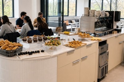 Buffet avec viennoiseries, fruits, brochettes et machine à café dans un espace lumineux avec des convives assis.