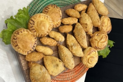 Plateau Africa (Pastels au thon, croustilles de manioc, feuilletés mafé poulet, feuilletés yassa poulet) Assortiment de petits chaussons salés et feuilletés disposés sur un plateau en bois avec des feuilles de salade.