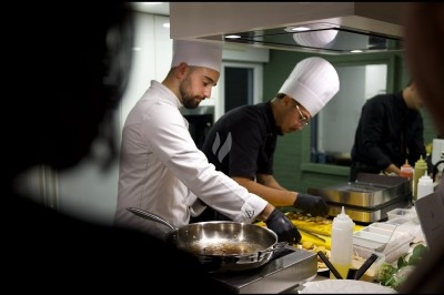 Deux chefs cuisiniers en tenue blanche préparent des plats dans une cuisine professionnelle éclairée.