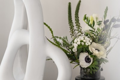 Sculpture blanche abstraite sur un socle noir, accompagnée d'un vase noir contenant un bouquet de fleurs blanches et vertes.