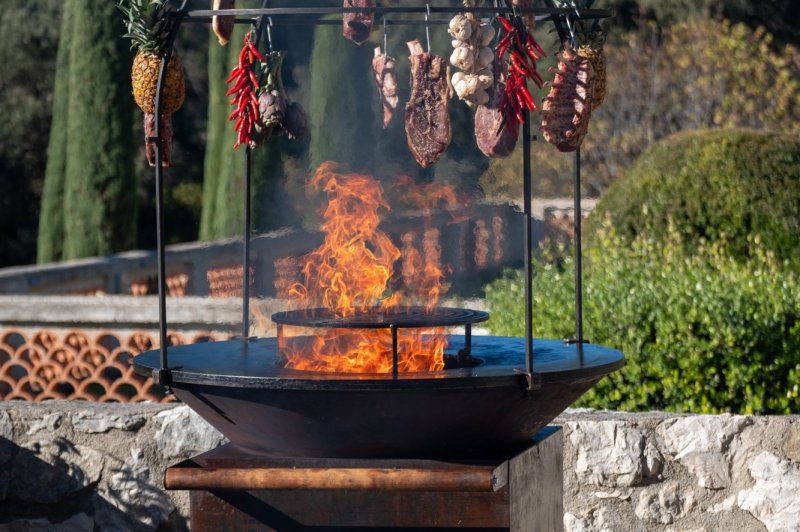 Barbecue extérieur avec feu vif, viandes, piments et ananas suspendus, entouré de végétation méditerranéenne.