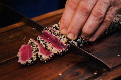 Main coupant un filet de thon mi-cuit en tranches, enrobé de graines de sésame, sur une planche en bois.