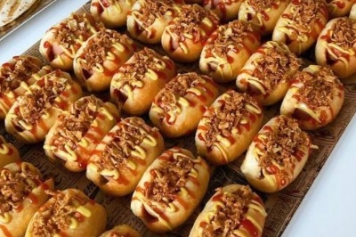 Rangées de petits pains garnis de sauce, moutarde et oignons frits, disposés sur un plateau.