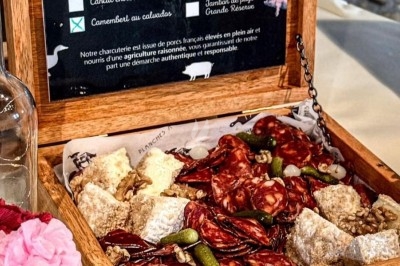 Plateau en bois ouvert contenant des charcuteries, fromages et cornichons, posé sur une table avec une fleur en décoration.