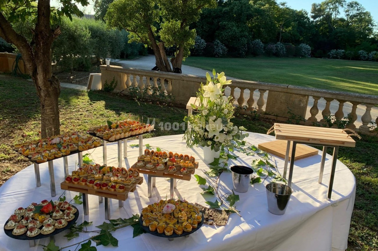 Buffet en extérieur avec des amuse-bouches variés disposés sur des présentoirs, entouré de verdure et de fleurs.