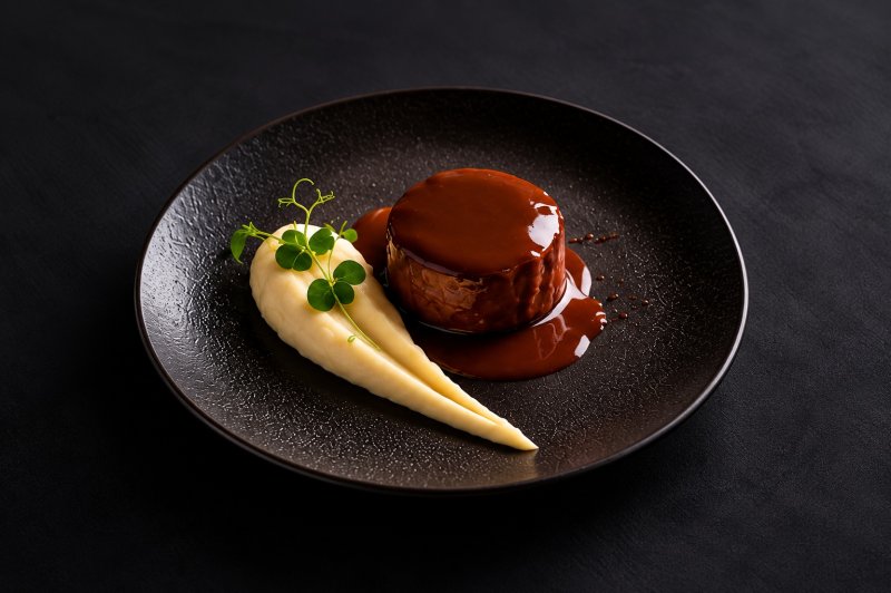 Assiette noire avec dessert chocolat brillant et accompagnement de crème pâle garni de microplante.