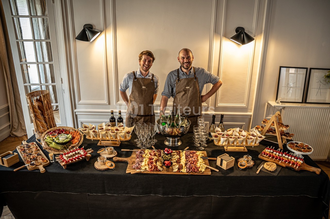 Deux serveurs en tablier présentent un buffet de charcuteries, fromages et canapés sur table noire en salle élégante.