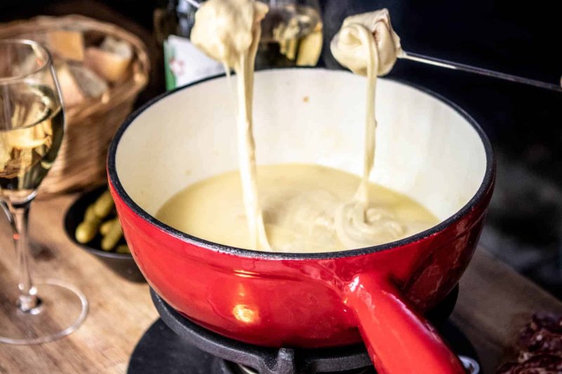 Caquelon à fondue savoyarde rouge et blanc avec fromage fondu et deux fourchettes, accompagné de verres de vin blanc.