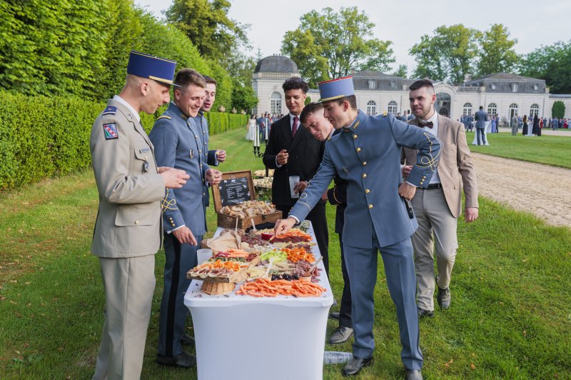 Militaires en uniforme autour d'une table de buffet froid en plein air, parc avec château en arrière-plan.